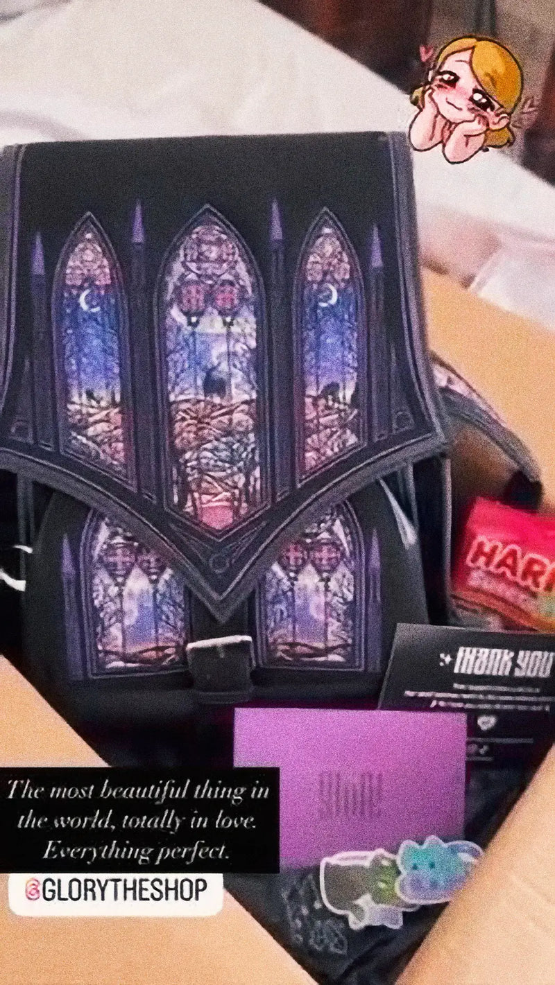 Unboxing of a black gothic backpack with purple stained glass window design, sitting in a cardboard box with Haribo sweets and a thank you card.