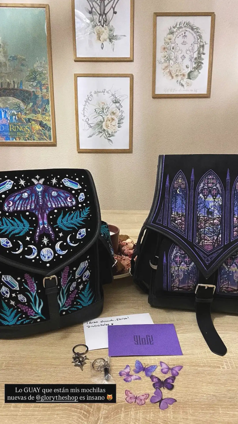 Dos mochilas góticas negras con bordados detallados de una polilla luna mística y ventanas de catedral en tonos púrpuras y azules sobre una mesa de madera.