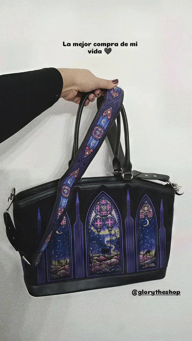 Woman holding a black gothic handbag featuring a purple stained glass cathedral design and a matching strap against a white wall.