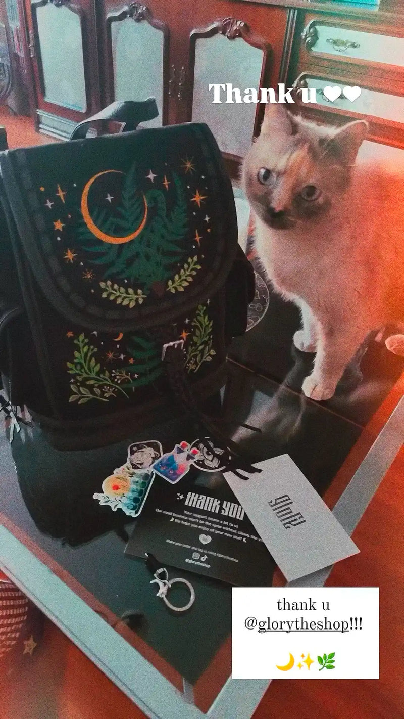 A light-colored cat sitting next to a black backpack embroidered with a green forest and crescent moon design on a table.