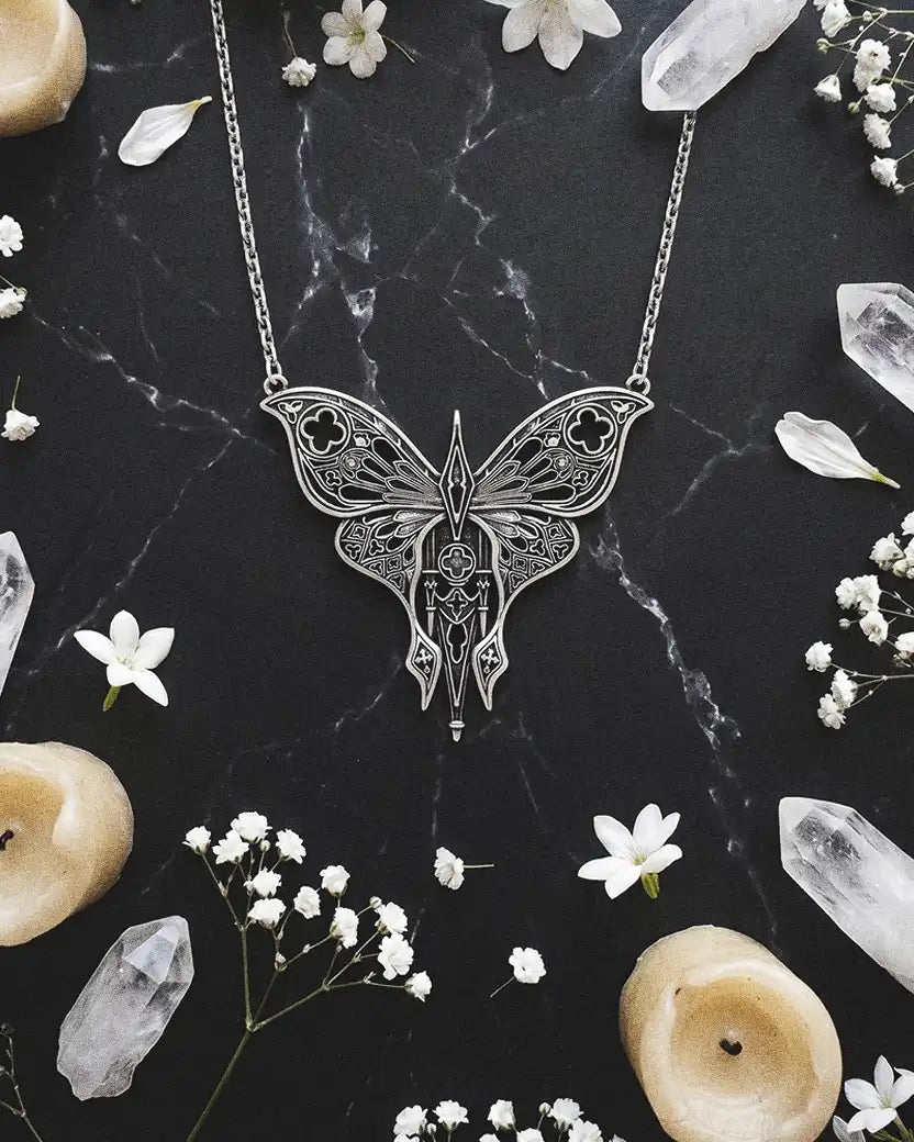 Imagen flat lay de un collar de la marca Restyle con forma de polilla apoyado sobre un fondo de mármol negro mate con vetas blancas, flores, cristales blancos y velas.