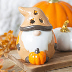Quemador de aceites esenciales con forma de gnomo adorable otoñal con calabaza y tapa en forma de gorro vista frontal sobre escritorio con decoración al rededor.