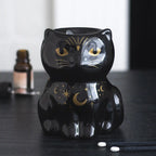 Quemador de aceites esenciales en forma de gato negro con estrellas en color dorado vista frontal sobre escritorio con decoración al rededor.
