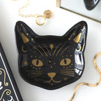 Plato con forma de gato en color negro con detalles en dorado apoyado en una mesa con joyería al rededor.