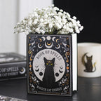Jarrón en forma de libro estilo witchy con título libro de hechizos y gato negro en la portada vista ángulo en escritorio con decoración al rededor. 