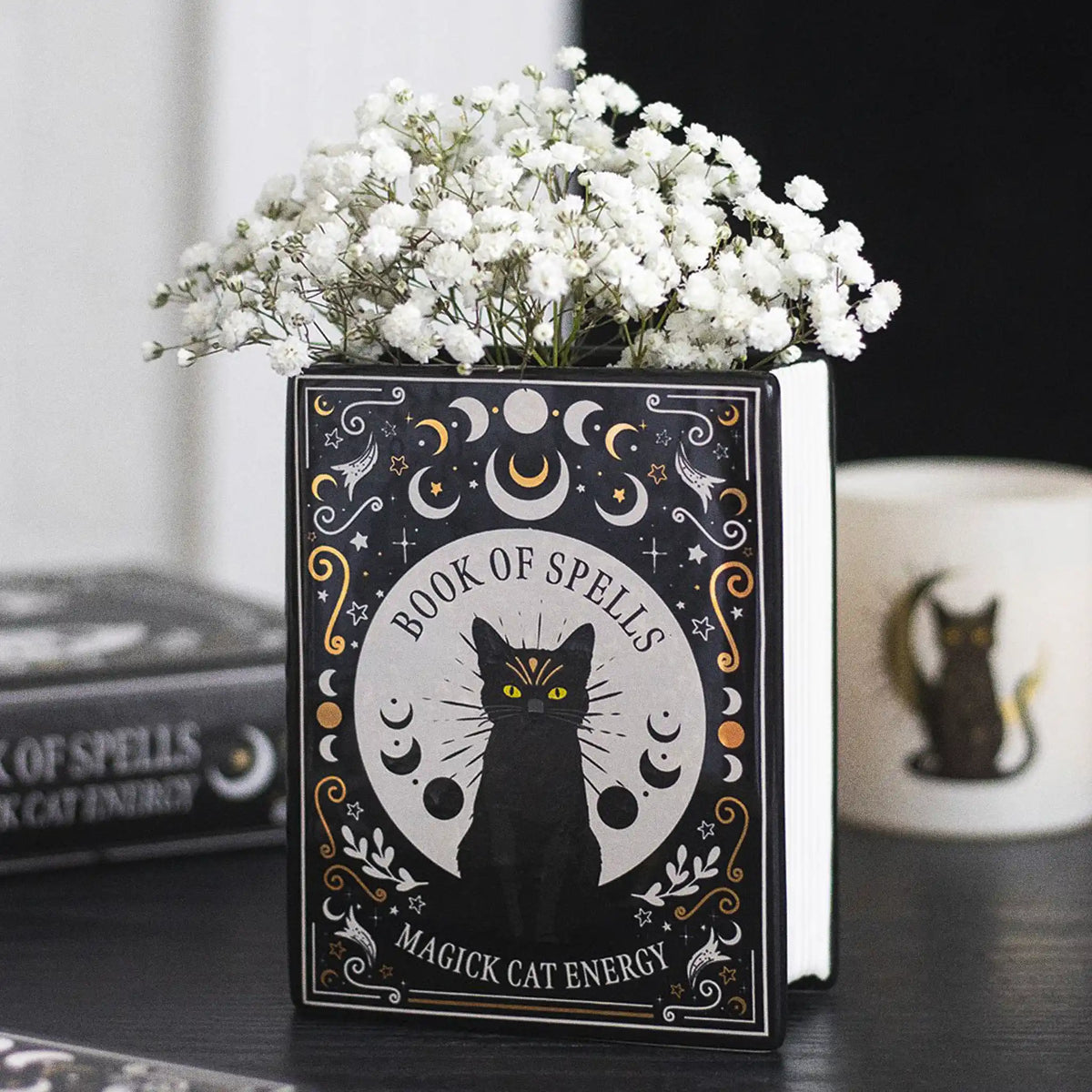 Jarrón en forma de libro estilo witchy con título libro de hechizos y gato negro en la portada vista ángulo en escritorio con decoración al rededor. 