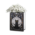 Jarrón en forma de libro estilo witchy con título libro de hechizos y gato negro en la portada vista sobre fondo blanco con flores.