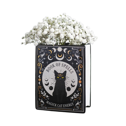 Jarrón en forma de libro estilo witchy con título libro de hechizos y gato negro en la portada vista sobre fondo blanco con flores.