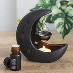 Quemador de aceite en forma de luna creciente con estrellas cut out en color negro con colgante metálico negro para poner aceite vista fontal sobre escritorio con decoración al rededor.