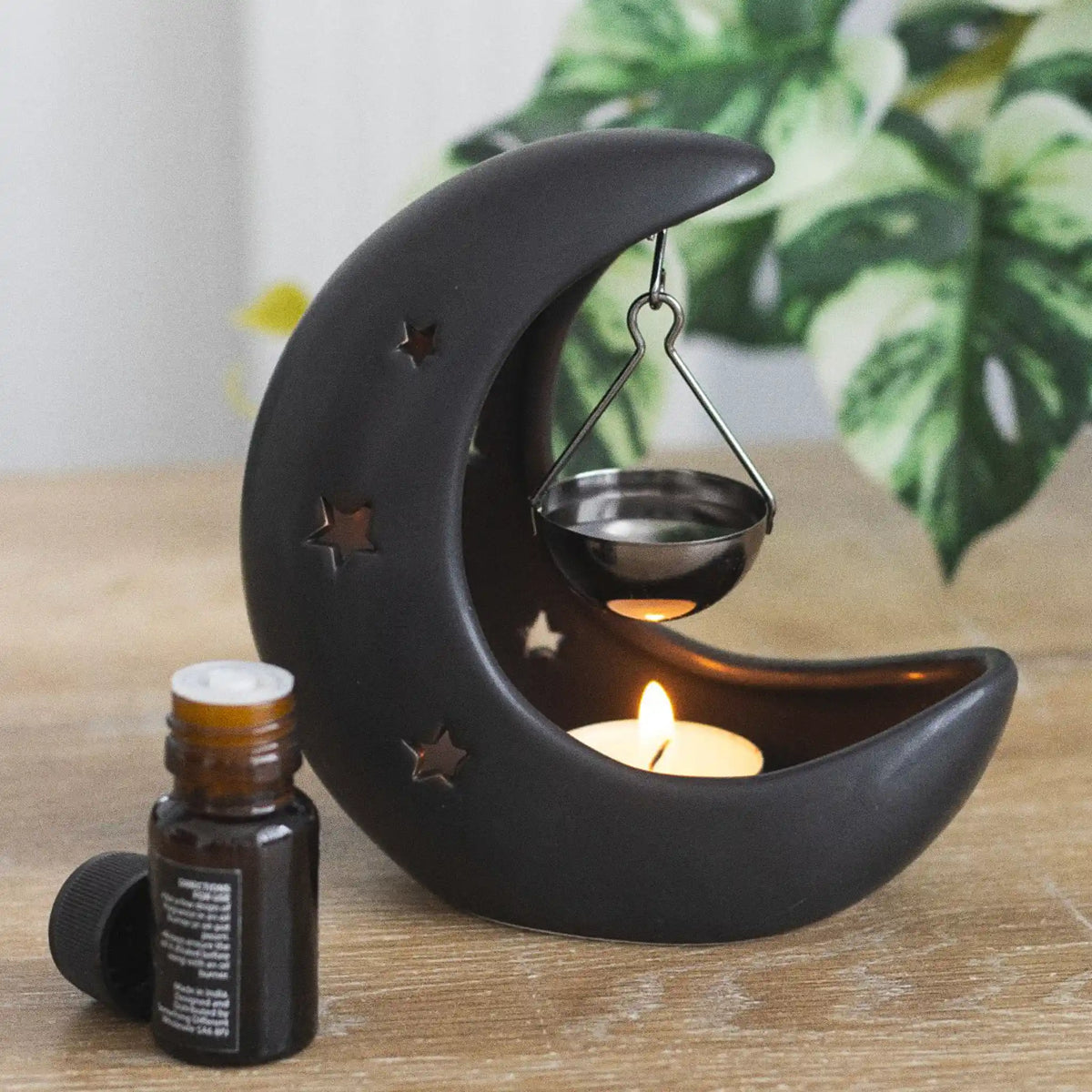 Quemador de aceite en forma de luna creciente con estrellas cut out en color negro con colgante metálico negro para poner aceite vista fontal sobre escritorio con decoración al rededor.