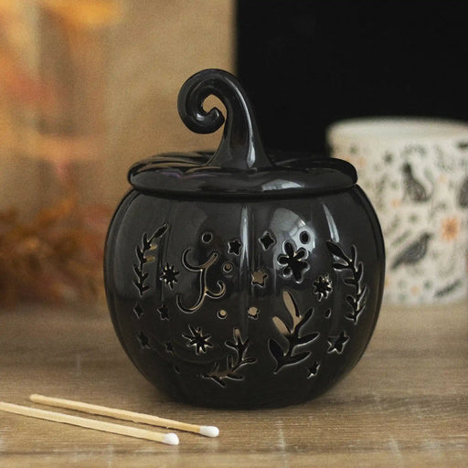 Quemador de aceites esenciales y calentador de cera en forma de calabaza con diseño calado otoñal perfecta para halloween y otoño en color negro vista frontal sobre escritorio con decoración al rededor.