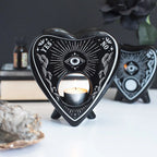 Quemador de aceites esenciales y calentador de cera estilo gótico de ouija en blanco y negro gótico vista frontal con vela encendida sobre escritorio con decoración al rededor.