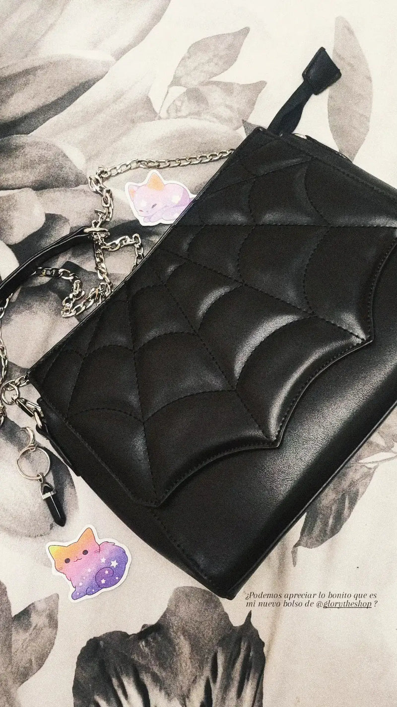 Aesthetic shot of a black spiderweb-shaped shoulder bag with a silver chain strap lying on a floral background with cat stickers.