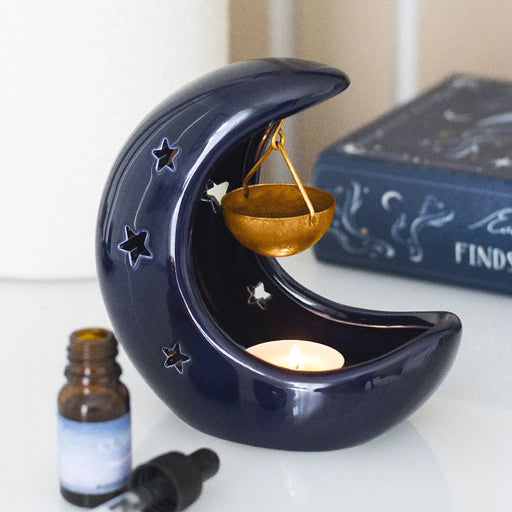 Quemador de aceites esenciales con forma de luna con estrellas cut out en color azul oscuro y colgante para aceites en metal dorado vista frontal sobre escritorio con decoración al rededor.