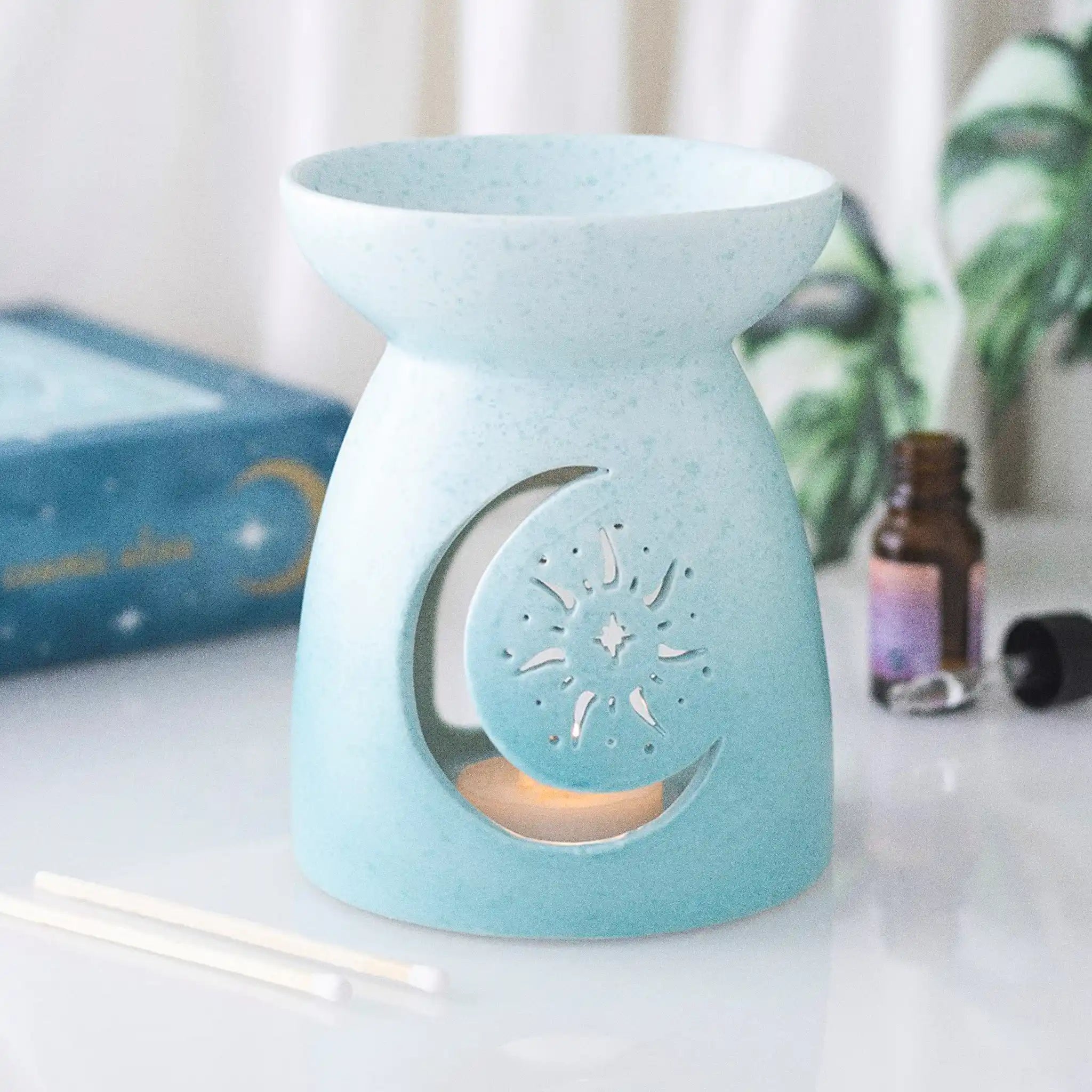 Quemador de aceites esenciales con forma clásica y diseño cut out en la parte frontal de luna y sol y color degradado en tonos azules vista sobre escritorio con decoración al rededor.