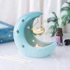 Quemador de aceites esenciales en forma de luna creciente con estrellas cut out en tono azul pastel con recipiente colgante vista frontal sobre escritorio con decoración al rededor.