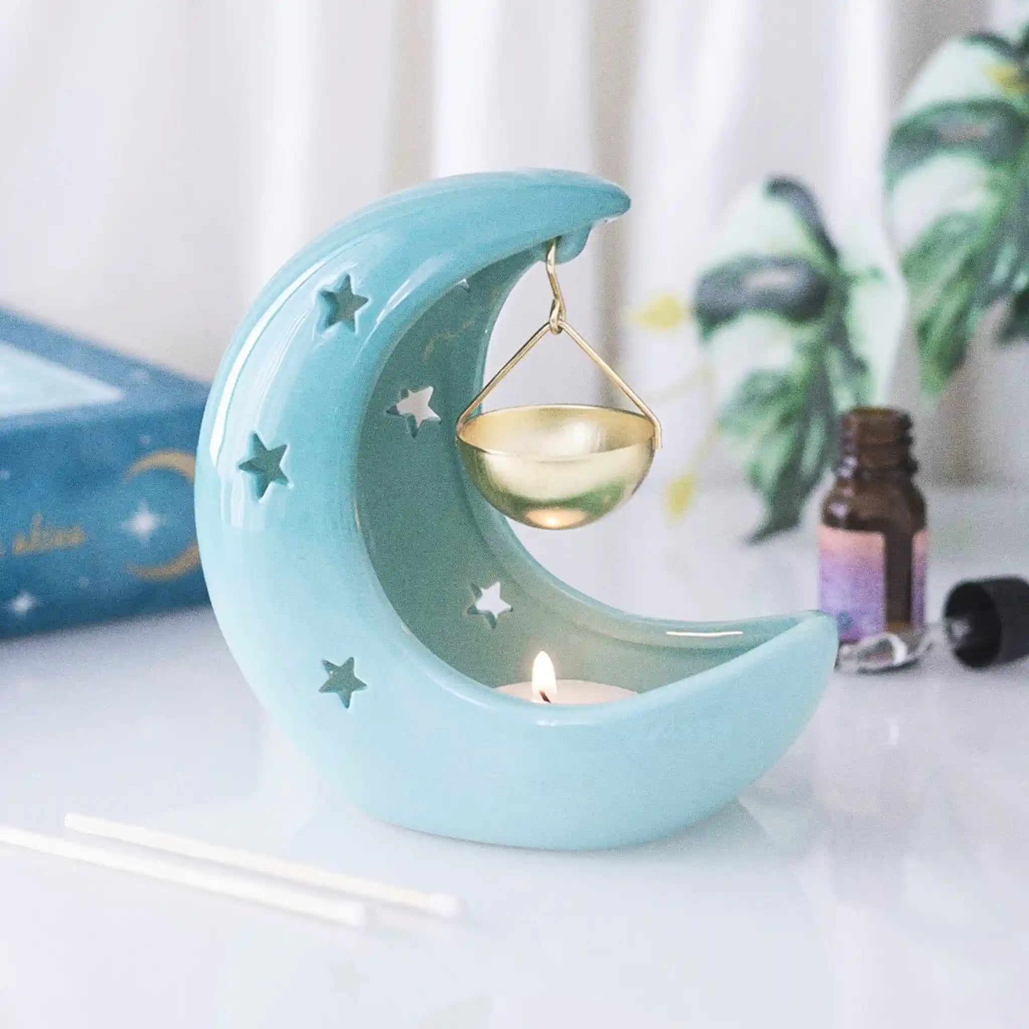 Quemador de aceites esenciales en forma de luna creciente con estrellas cut out en tono azul pastel con recipiente colgante vista frontal sobre escritorio con decoración al rededor.