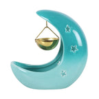 Quemador de aceites esenciales en forma de luna creciente con estrellas cut out en tono azul pastel con recipiente colgante vista posterior sobre fondo blanco.