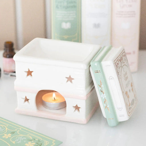 Quemador de aceites esenciales y cera en forma de libros apilados estilo fairy aesthetic vista de parte posterior sin la tapa con vela encendida sobre escritorio con decoración al rededor.
