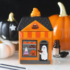 Quemador de aceites esenciales con forma de mini panadería fantasmal estética de Halloween para el hogar vista frontal sobre escritorio con decoración al rededor.