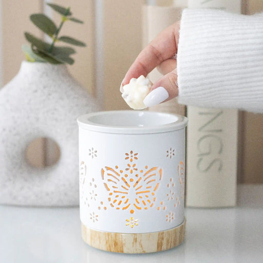 Quemador de aceites esenciales y calentador de cera con diseño de mariposa calada en blanco y base de madera vista frontal sobre escritorio con decoración al rededor.
