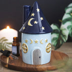 Quemador de aceites esenciales con decoración estilo whimsical y celestial de estrellas y lunas en pequeña cabaña acogedora vista frontal sobre escritorio con decoración al rededor.