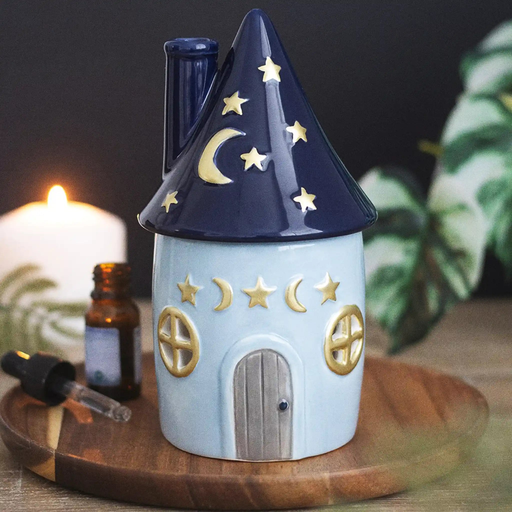 Quemador de aceites esenciales con decoración estilo whimsical y celestial de estrellas y lunas en pequeña cabaña acogedora vista frontal sobre escritorio con decoración al rededor.