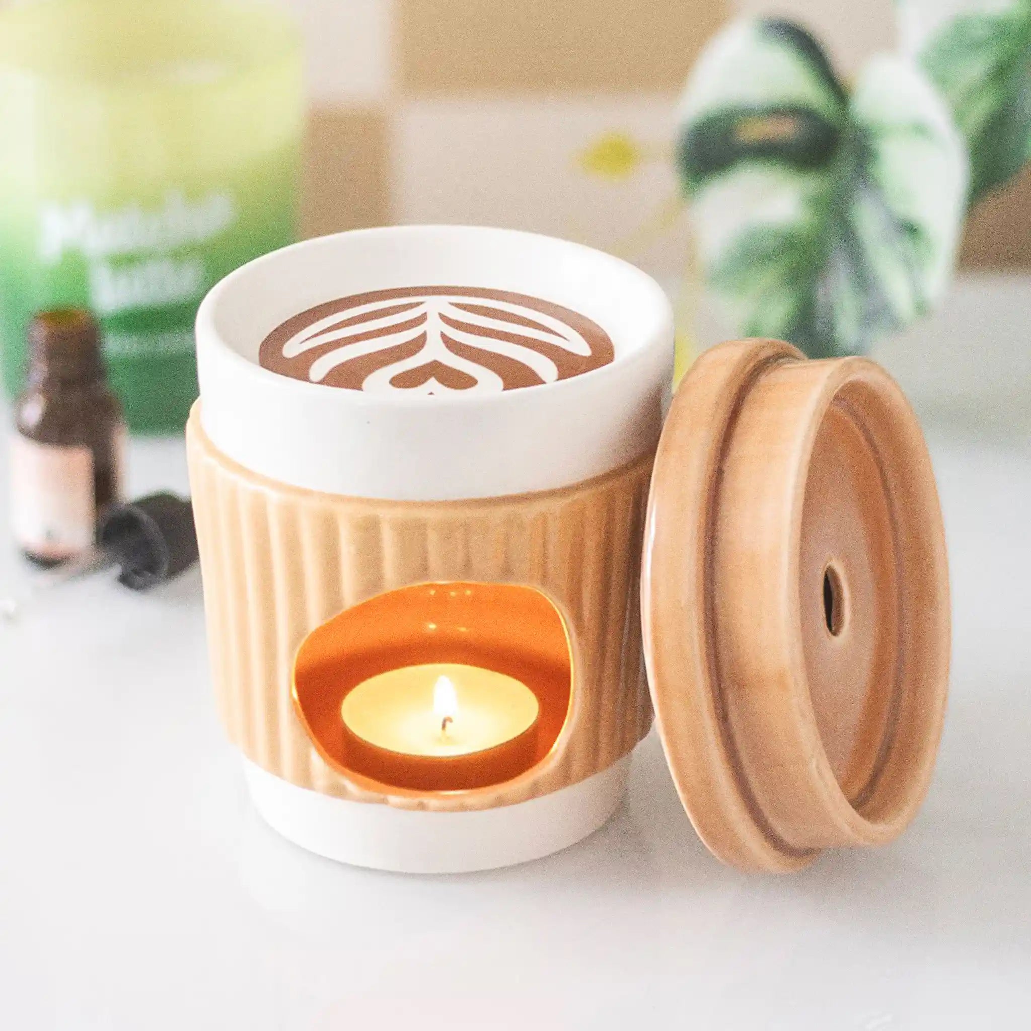 Quemador de aceites esenciales con forma de taza de café take away aesthetic vista posterior con tapa quitada del hueco para la vela de te sobre escritorio con decoración al rededor.