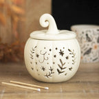 Quemador de aceites esenciales y calentador de cera con diseño de calabaza calado con motivos otoñales en blanco roto vista frontal sobre escritorio con decoración al rededor.