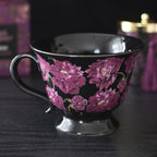 Taza negra de estética dark romance en negro con flores en color púrpura vista de taza y asa sobre escritorio con decoración al rededor.