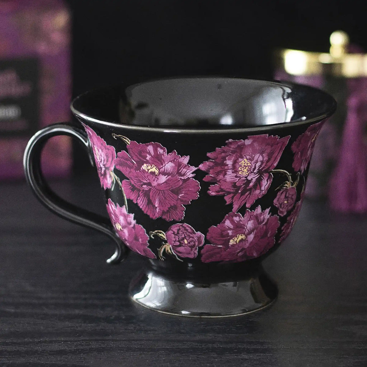 Taza negra de estética dark romance en negro con flores en color púrpura vista de taza y asa sobre escritorio con decoración al rededor.