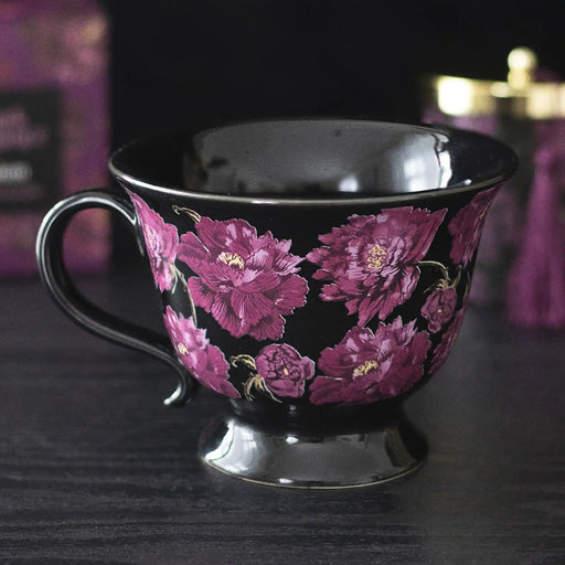 Taza negra de estética dark romance en negro con flores en color púrpura vista de taza y asa sobre escritorio con decoración al rededor.