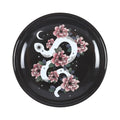 Plato de joyería redondo negro con diseño de serpiente enroscada en flores, estrellas y luna vista desde arriba sobre fondo blanco.
