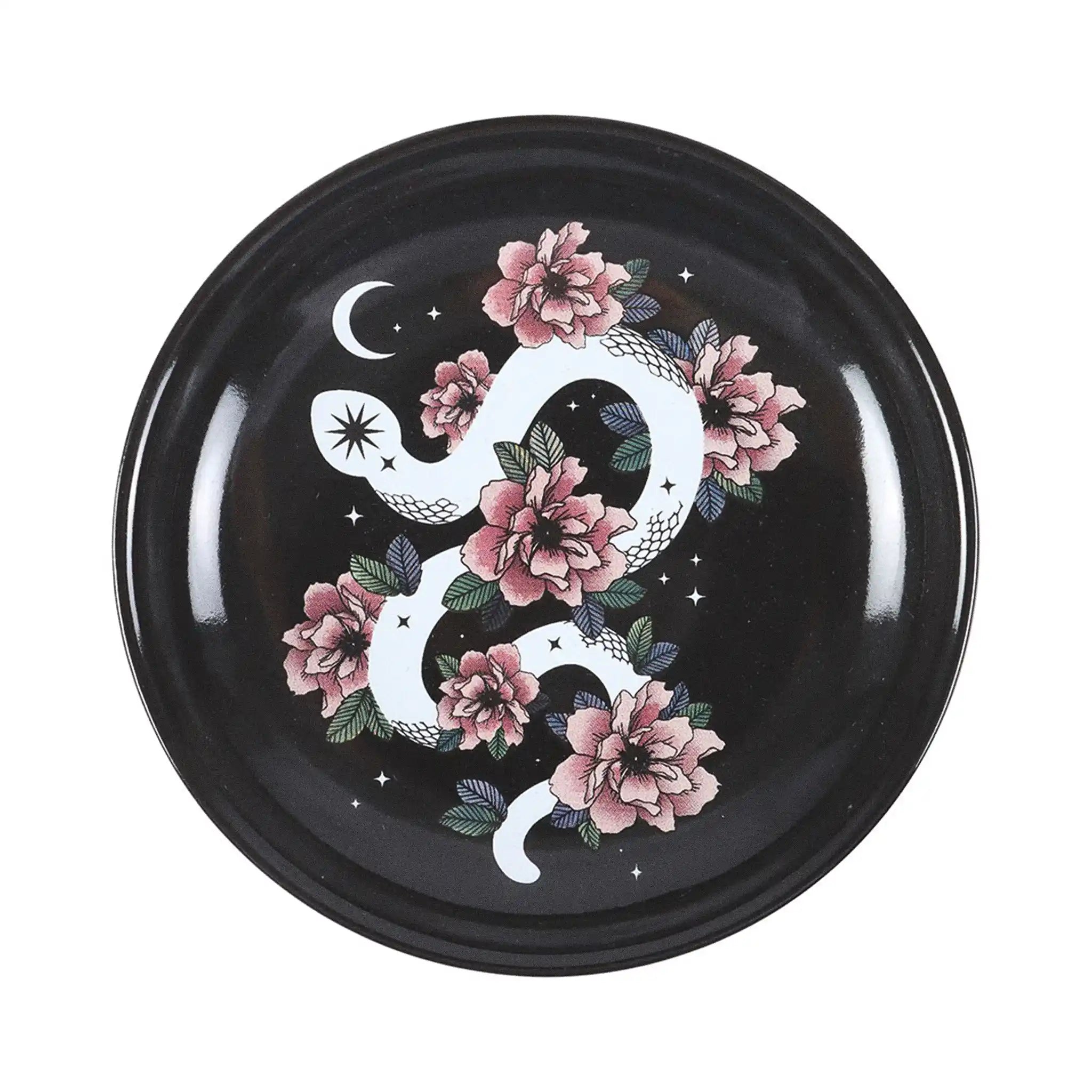 Plato de joyería redondo negro con diseño de serpiente enroscada en flores, estrellas y luna vista desde arriba sobre fondo blanco.