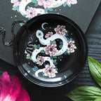 Plato de joyería redondo negro con diseño de serpiente enroscada en flores, estrellas y luna vista desde arriba sobre escritorio con joyería al rededor. 