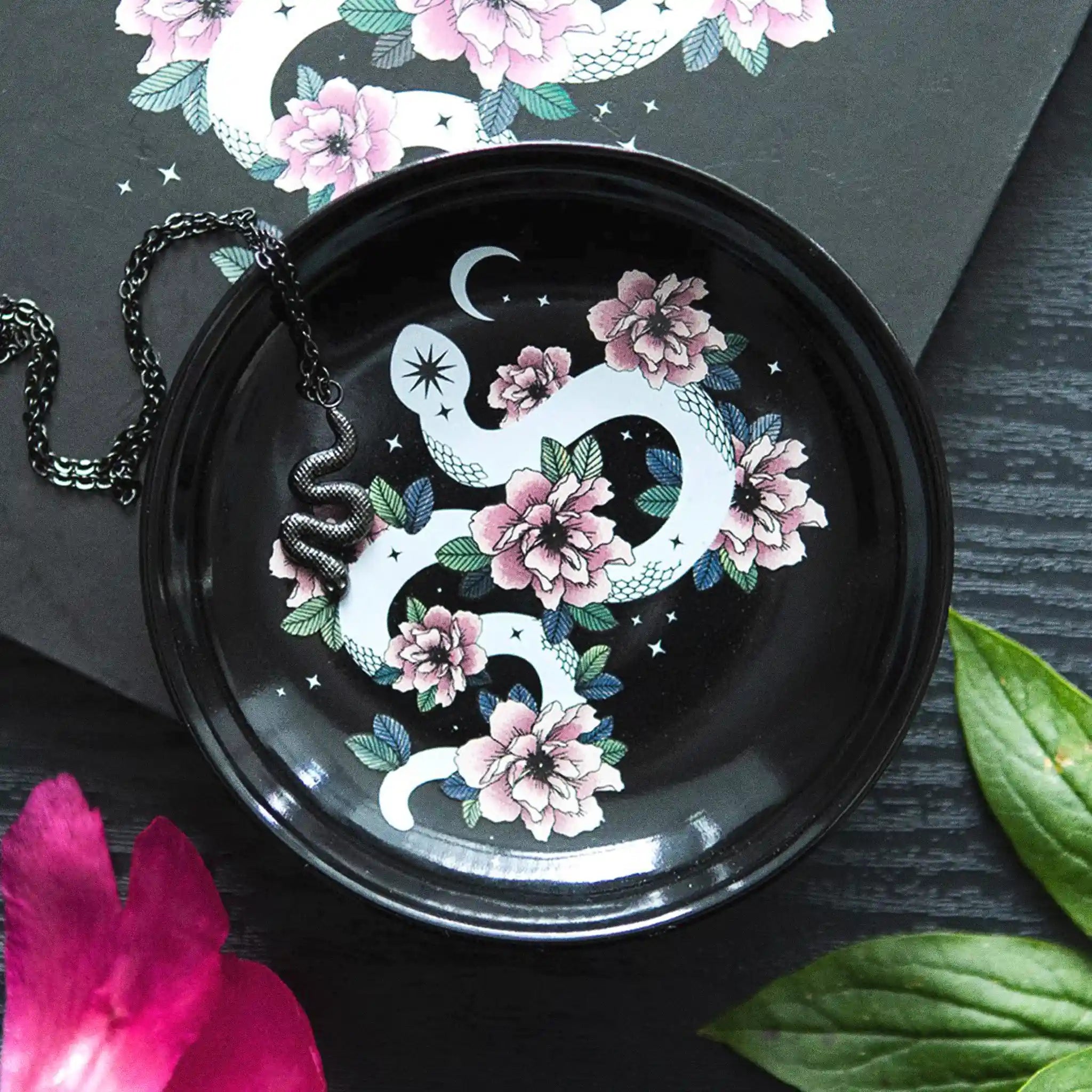 Plato de joyería redondo negro con diseño de serpiente enroscada en flores, estrellas y luna vista desde arriba sobre escritorio con joyería al rededor. 