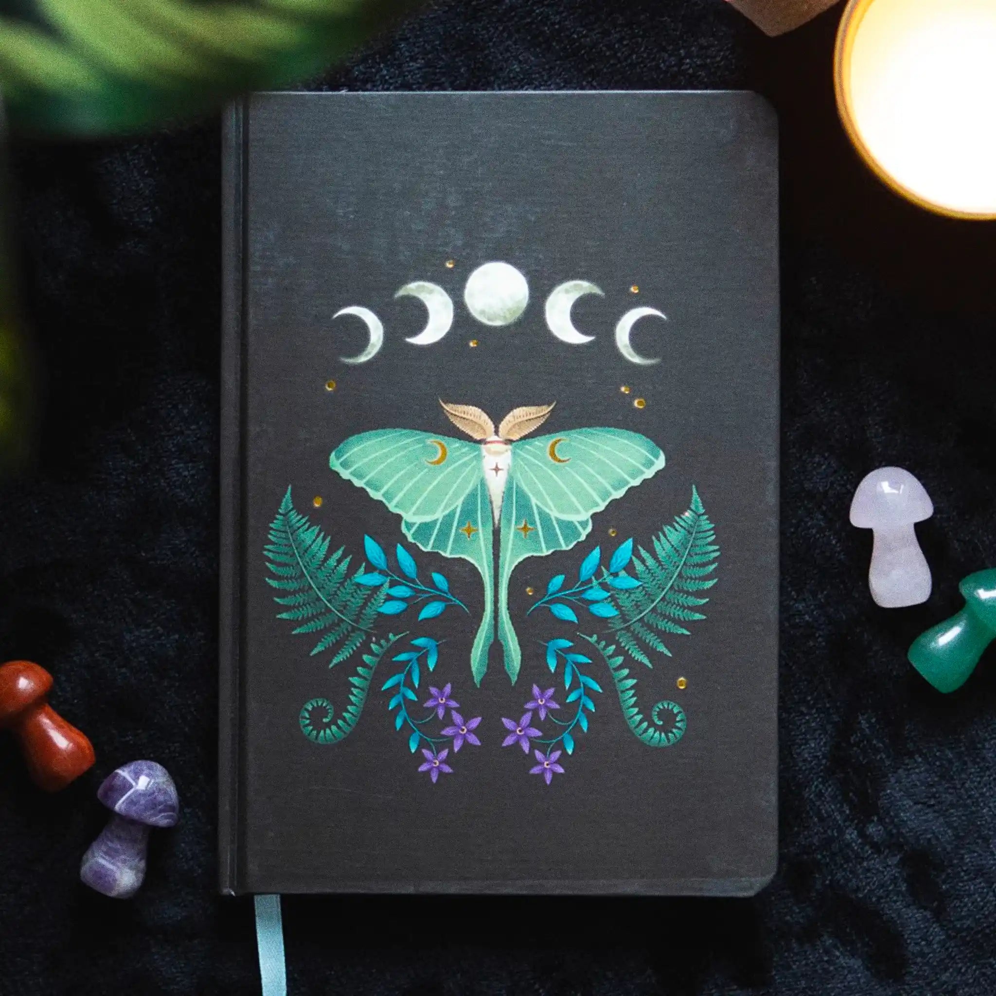 Libreta a5 apoyada sobre una manta con ilustración de lunas, polilla y helechos.