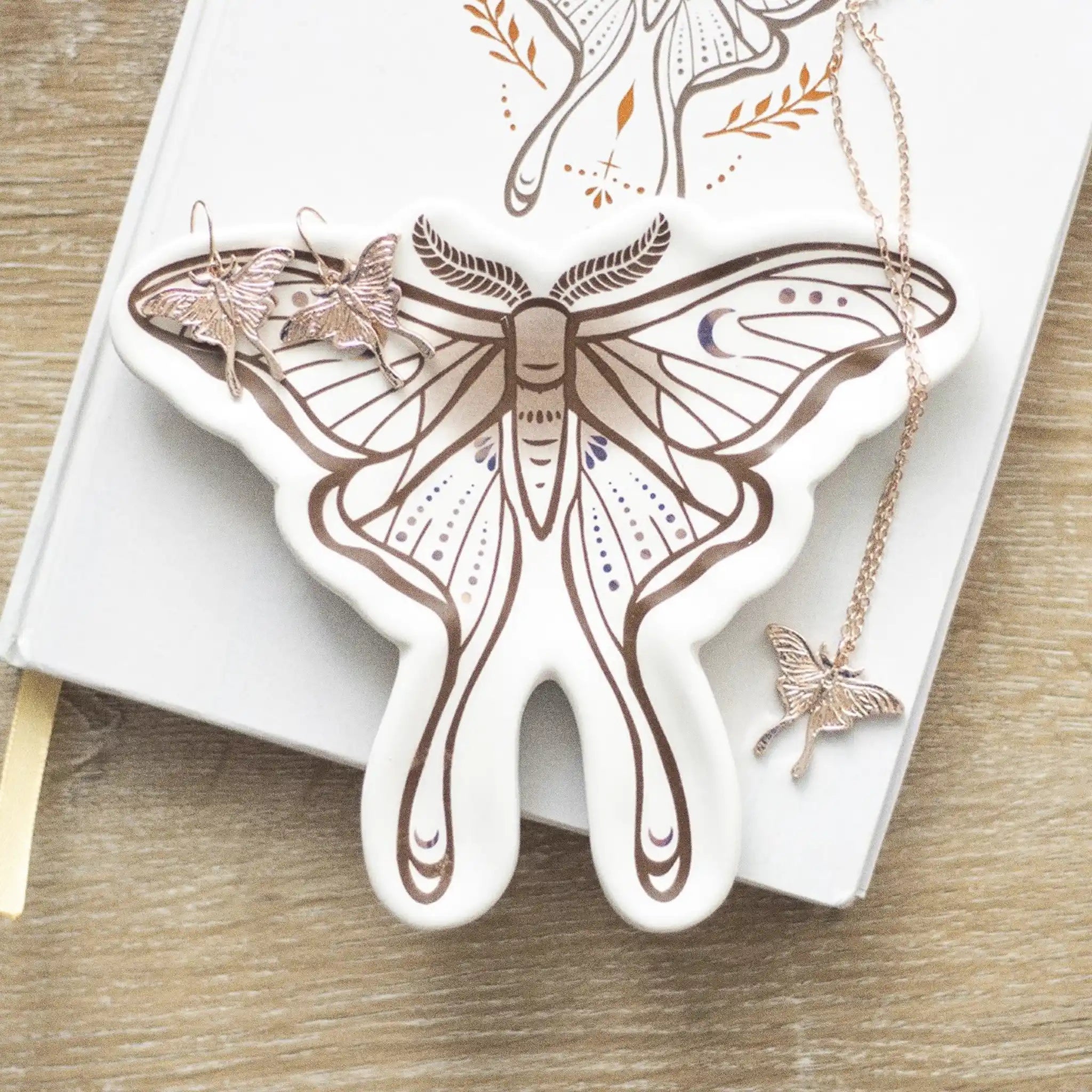 Bandeja para joyería con forma de polilla en blanco con estampado en tonos marrones sobre escritorio con joyería al rededor.