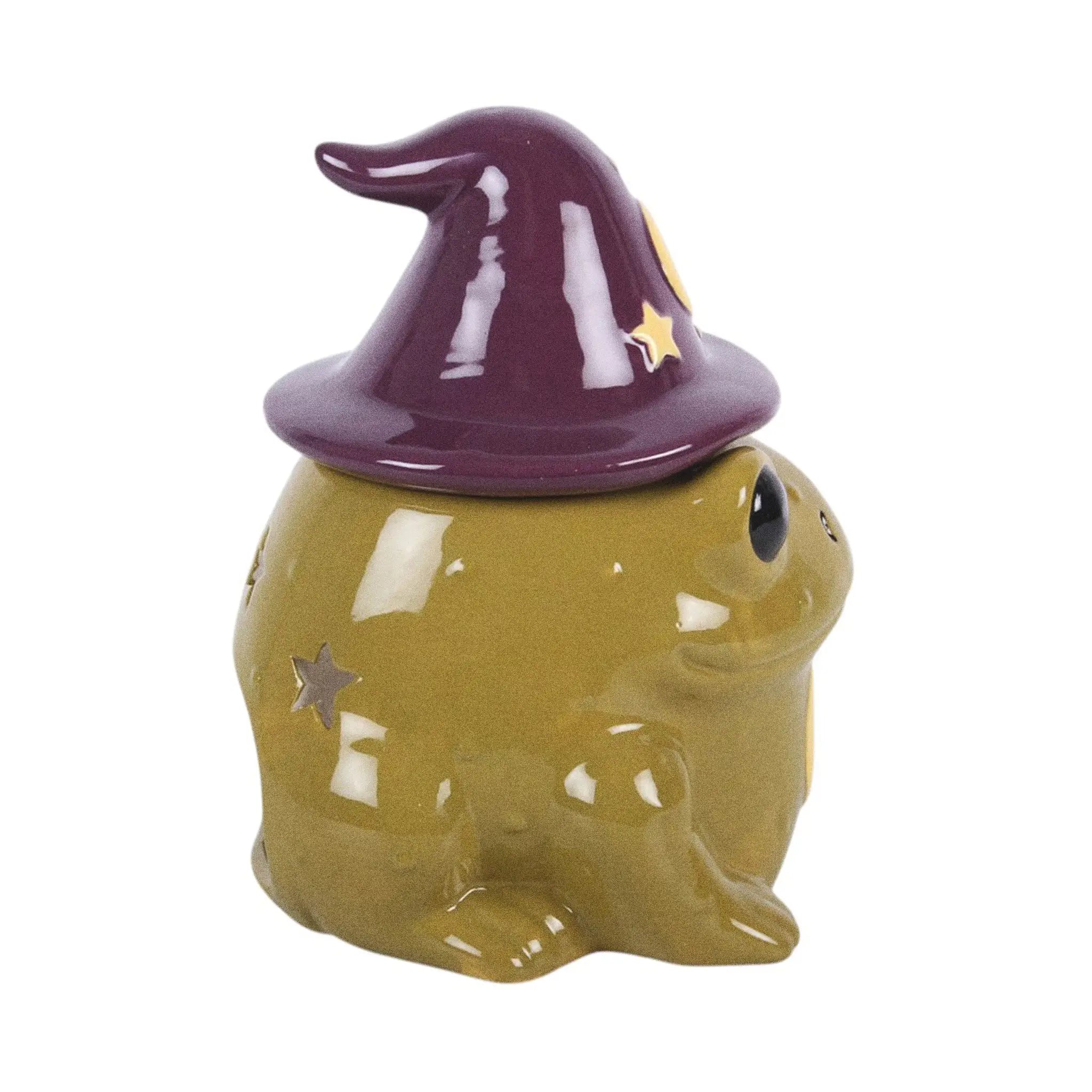 Quemador de aceites esenciales y calentador de cera en forma de sapo mágico con gorro de brujo con estrellas y luna estilo whimsical vista lateral sobre fondo blanco.