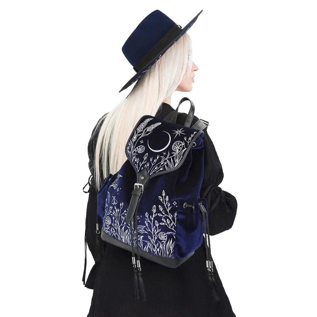Mochila gótica estilo whimsigoth en tejido de terciopelo combinado con piel sintética con bordados en plateado de luna, flores y estrellas y borlas decorativas vista en hombro de modelo