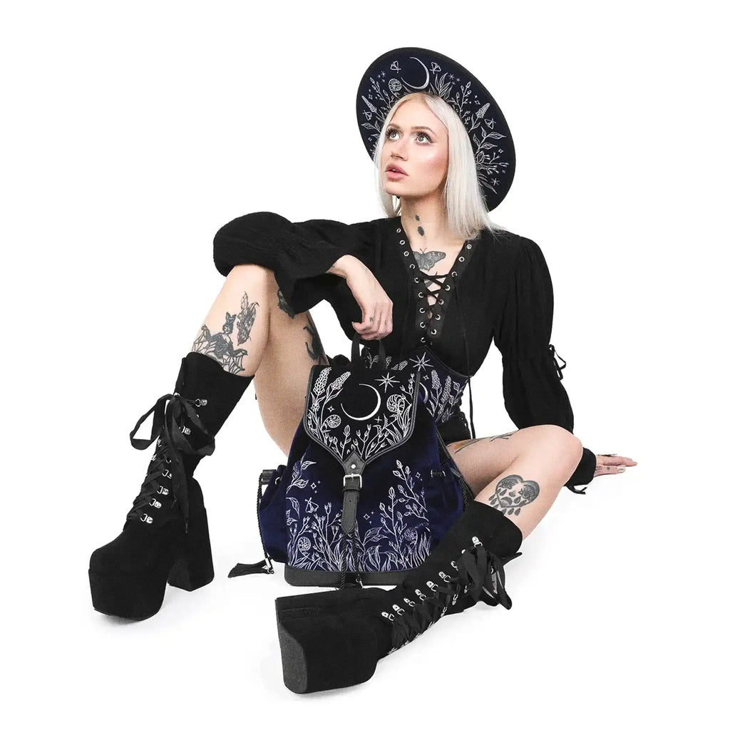 Mochila gótica estilo whimsigoth en tejido de terciopelo combinado con piel sintética con bordados en plateado de luna, flores y estrellas y borlas decorativas vista con modelo sentada