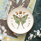 Plato joyero redondo con diseño de polilla verde y marrón y fases lunares en acabado metalizado dorado vista desde arriba sobre escritorio con decoración al rededor.