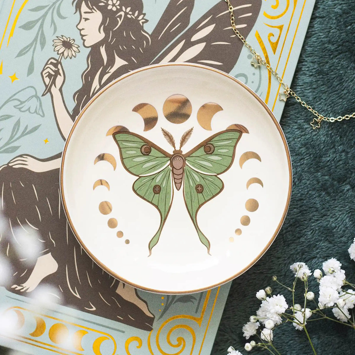Plato joyero redondo con diseño de polilla verde y marrón y fases lunares en acabado metalizado dorado vista desde arriba sobre escritorio con decoración al rededor.