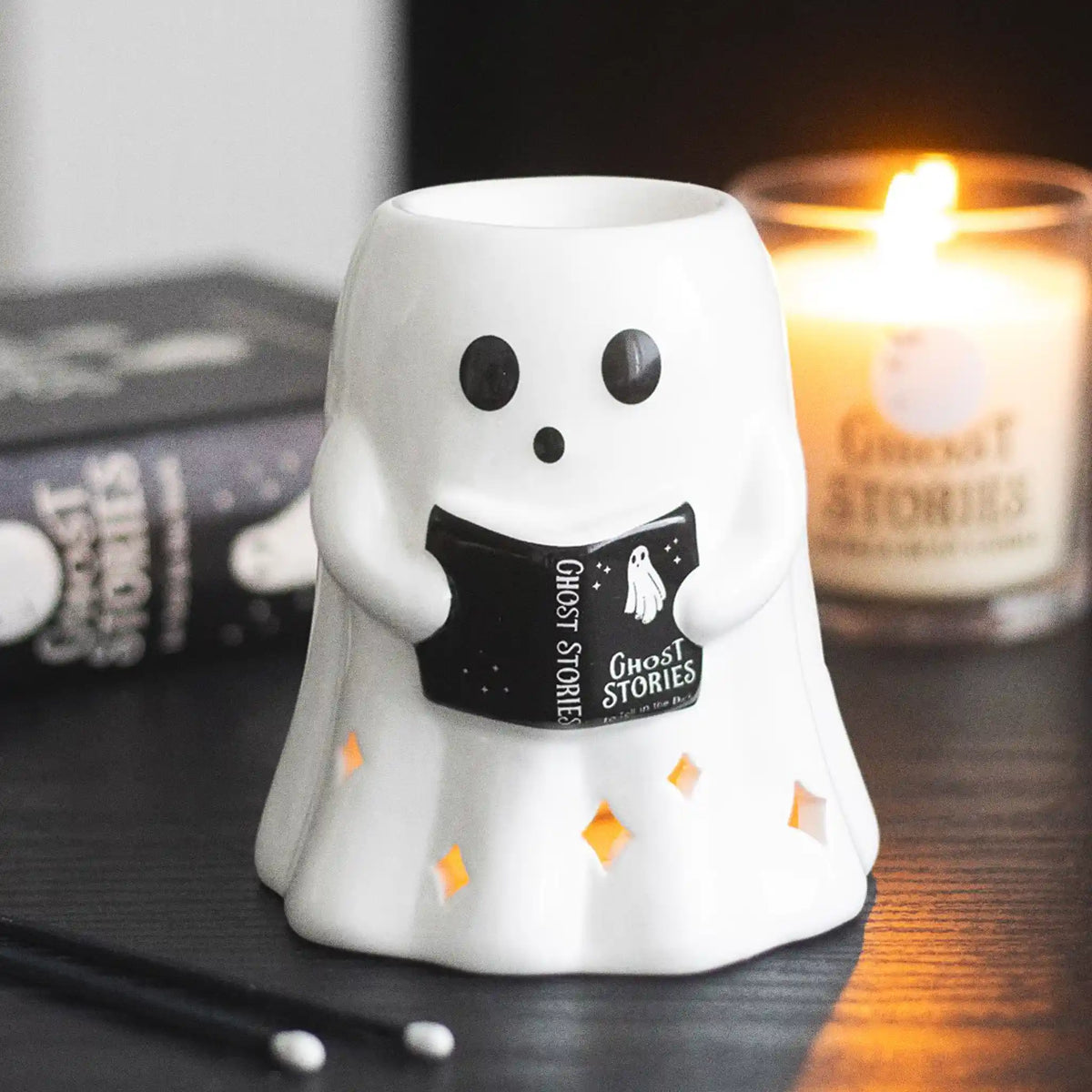 Quemador de aceites esenciales en forma de fantasma leyendo un libro estilo spooky halloween vista frontal sobre escritorio con decoración al rededor.