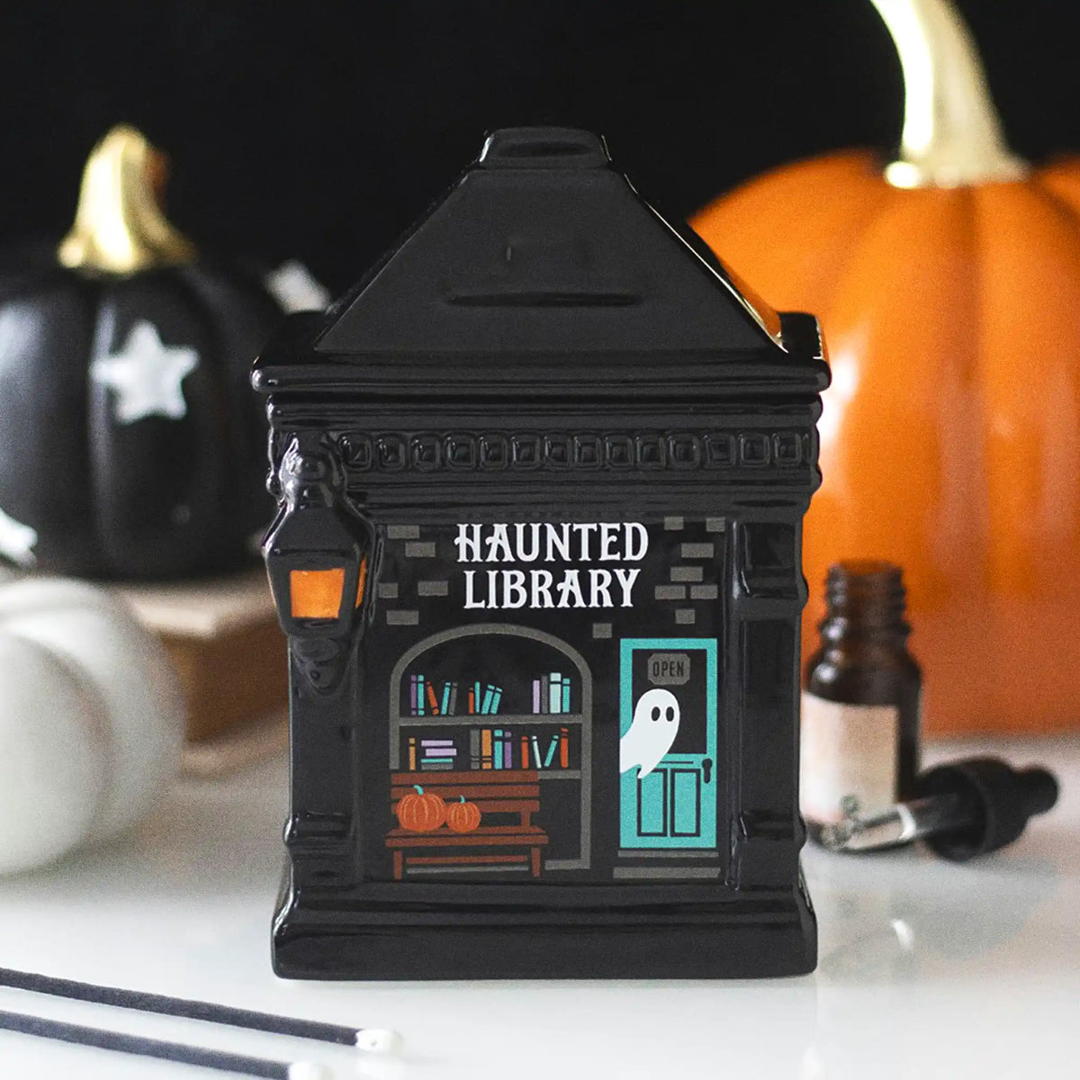Quemador de aceite y calentador de cera con forma de librería encantada estilo spooky halloween vista de frente sobre escritorio con decoración al rededor.