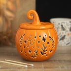 Quemador de aceites esenciales y calentador de cera con diseño naranja en forma de calabaza con motivos otoñales calados en la parte frontal vista frontal sobre escritorio con decoración al rededor.