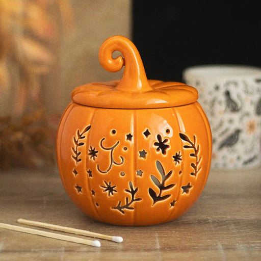 Quemador de aceites esenciales y calentador de cera con diseño naranja en forma de calabaza con motivos otoñales calados en la parte frontal vista frontal sobre escritorio con decoración al rededor.