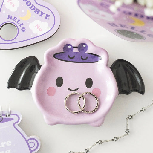 Plato joyero en forma de murciélago con alas estilo kawaii en tonos rosados con carita sonriente vista desde arriba sobre escritorio con decoración al rededor.