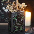 Jarrón en forma de libro estética gótica con título "Library of Poisonous Plants" aesthetic vista sobre escritorio con decoración al rededor y flores.