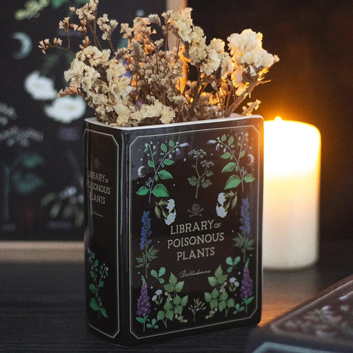 Jarrón en forma de libro estética gótica con título "Library of Poisonous Plants" aesthetic vista sobre escritorio con decoración al rededor y flores.